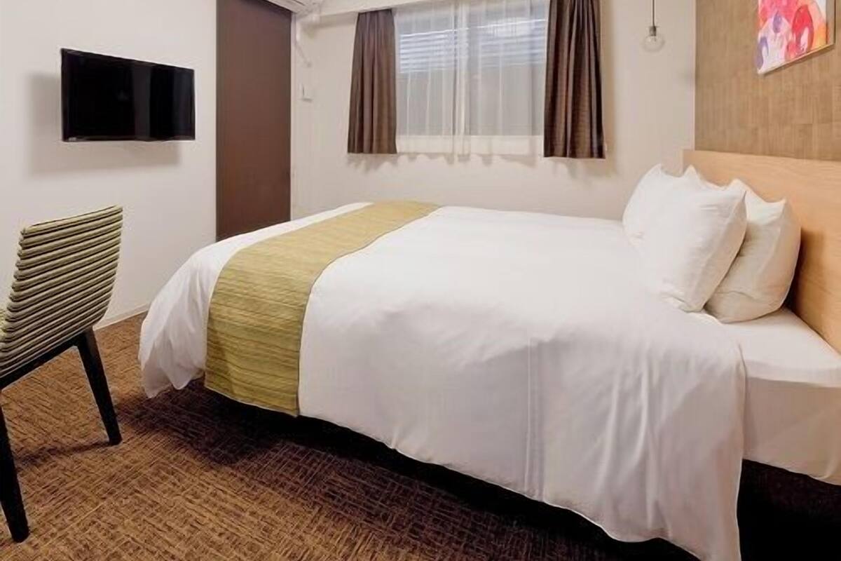 A hotel room with a bed, a chair, a flat-screen TV, an air conditioning unit, a window with curtains, and a painting on the wall.