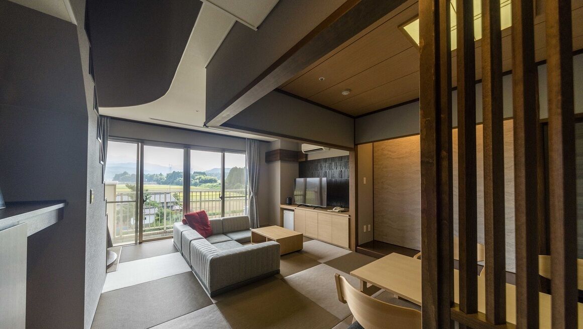 A modern hotel room with a balcony, a sofa, a dining table, and a kitchen area.