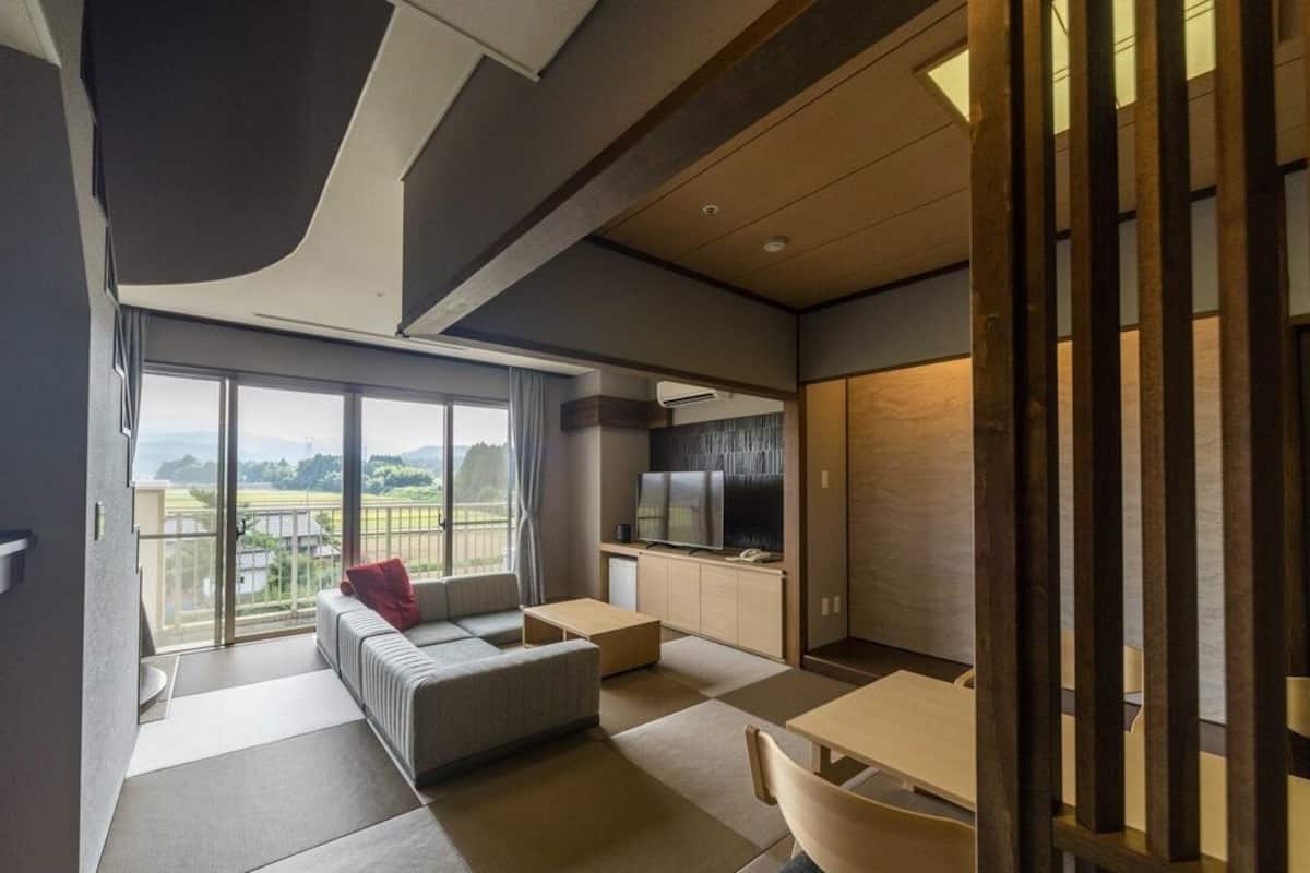 A modern hotel room with a balcony, a sofa, a dining table, and a kitchen area.