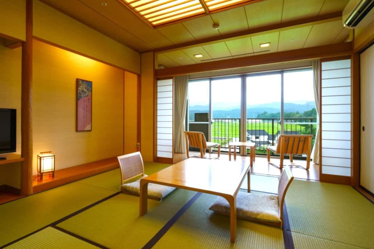 Una habitación tradicional japonesa con suelo de tatami, televisor, una mesa baja y vistas a un paisaje verde a través de amplios ventanales.