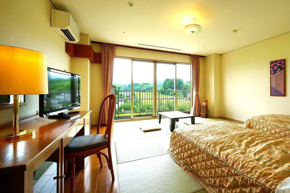 A hotel room with a bed, a desk with a TV, a chair, a balcony with a view, and a painting on the wall.