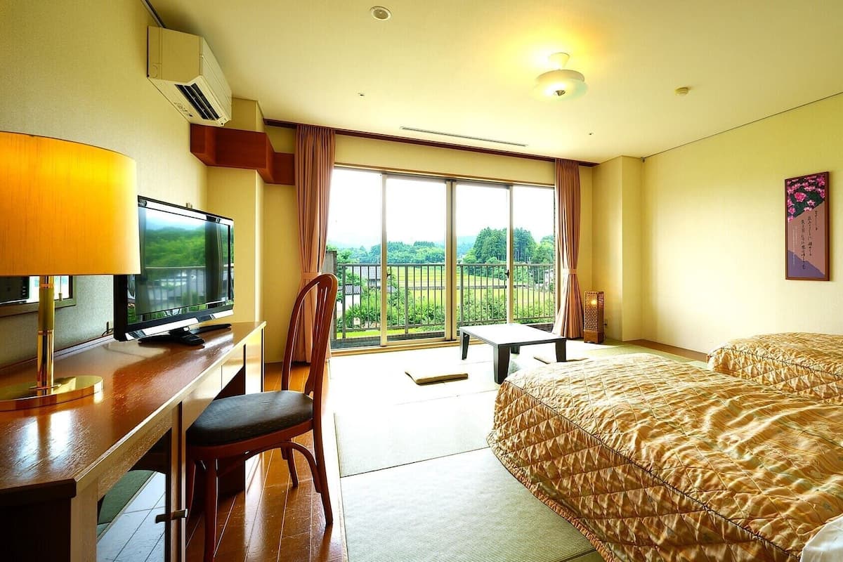 Habitación de hotel con cama, escritorio con televisor, silla, balcón con vistas y un cuadro en la pared.