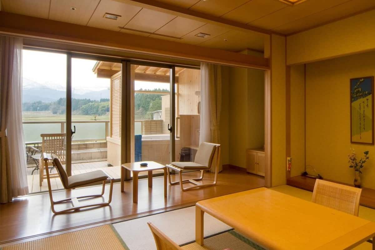 A room with a sliding glass door leading to a balcony with a view of a lake and mountains. There are two chairs and a small table on the balcony, and a larger table and chairs inside the room.