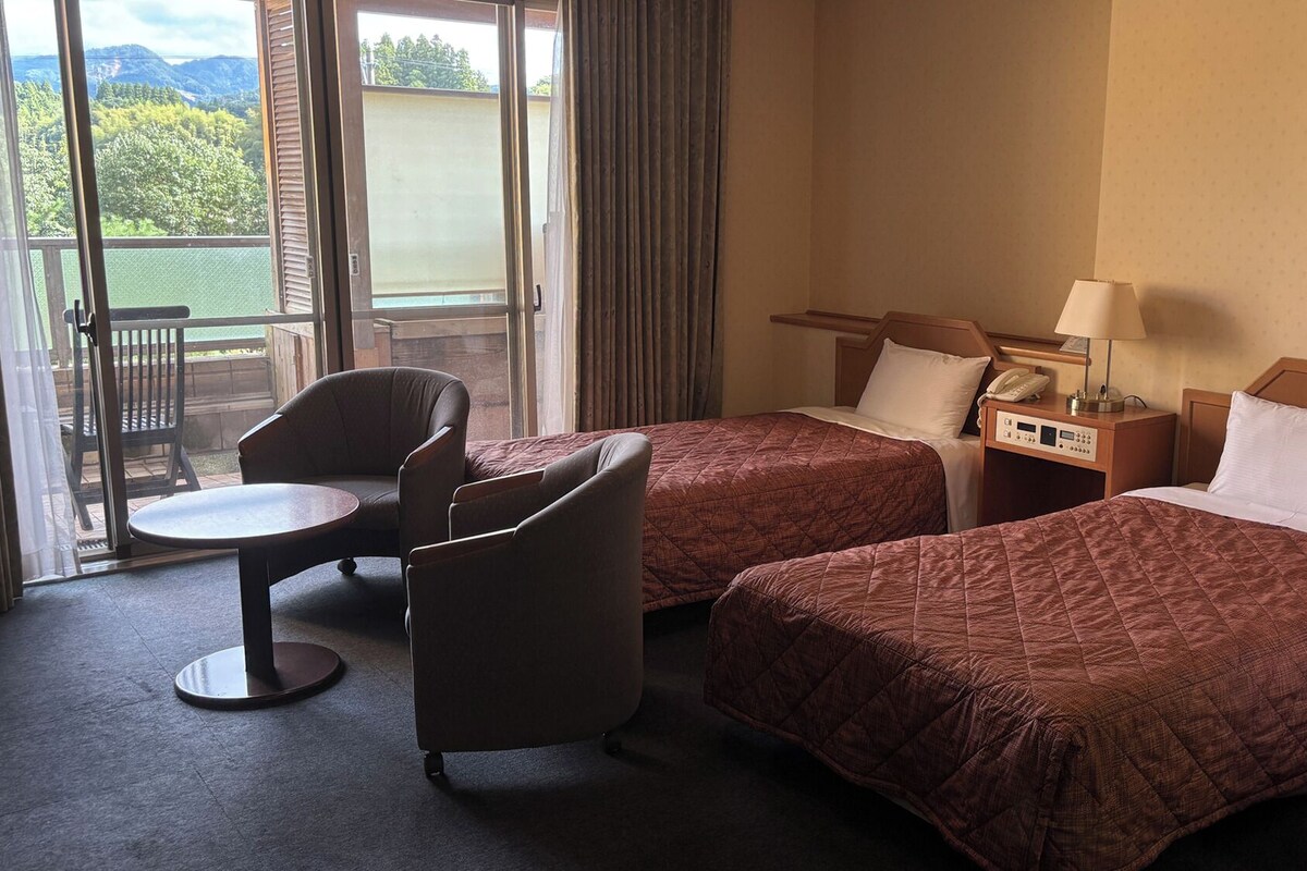 A hotel room with two beds, a small table, a chair, and a view of the outdoors.