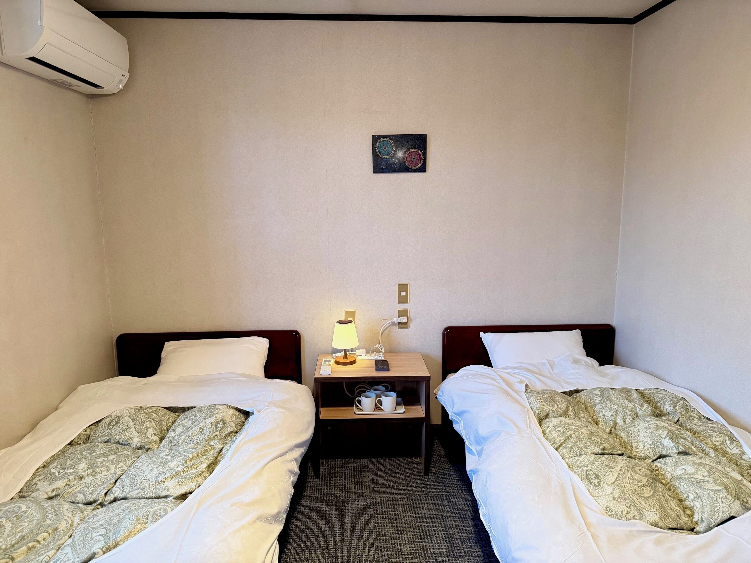A hotel room with two single beds, a bedside table with a lamp, a small painting on the wall, and an air conditioning unit.