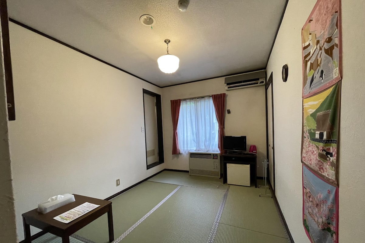 A room with tatami mat flooring, a desk with a computer, a TV, and a wall-mounted air conditioner.