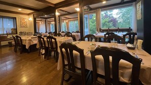A restaurant with multiple tables, wooden chairs, and large windows offering a view of greenery. - Hotel Sejour mint (Hakuba)