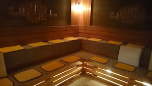 A sauna with wooden benches and benches with yellow mats, a wall-mounted light, and a tiled floor. - Hiroshima Capsule Hotel & Sauna New Japan EX - Caters to Men (Hiroshima)