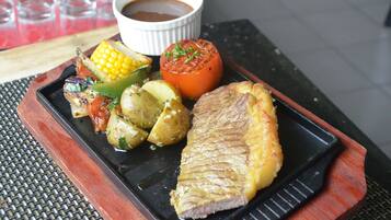 A black plate with grilled meat, corn on the cob, potatoes, and a tomato, accompanied by a small bowl of sauce.