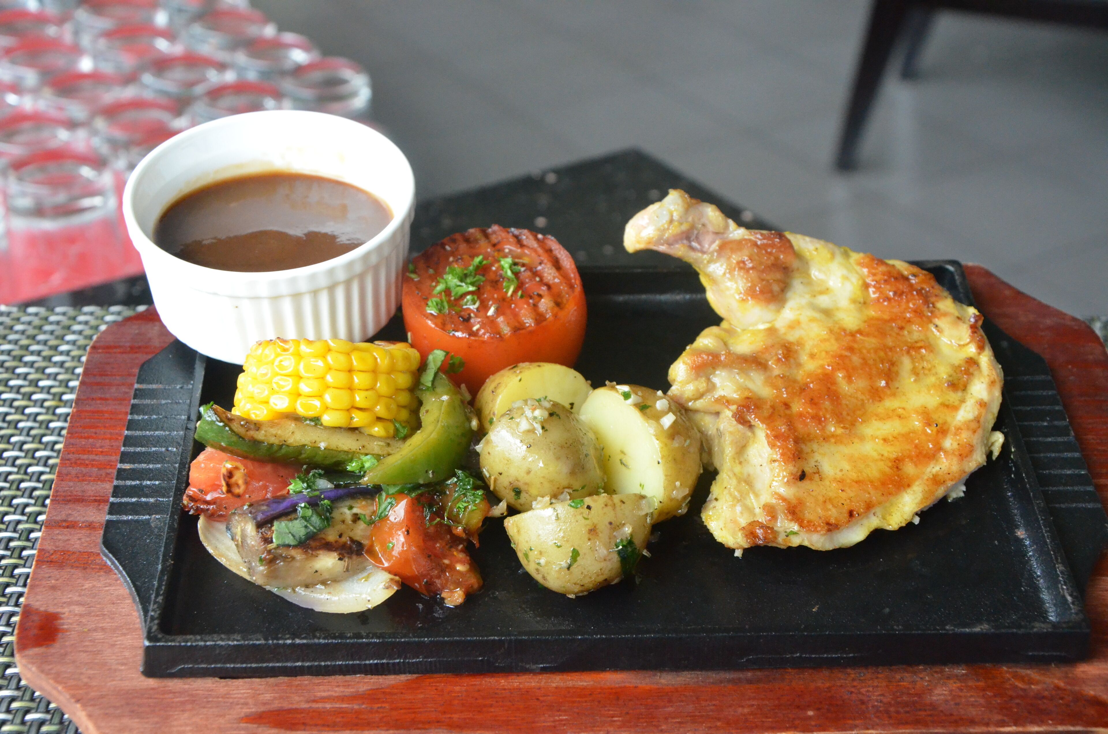 A black plate with grilled chicken, corn, potatoes, and a tomato, accompanied by a small bowl of sauce.