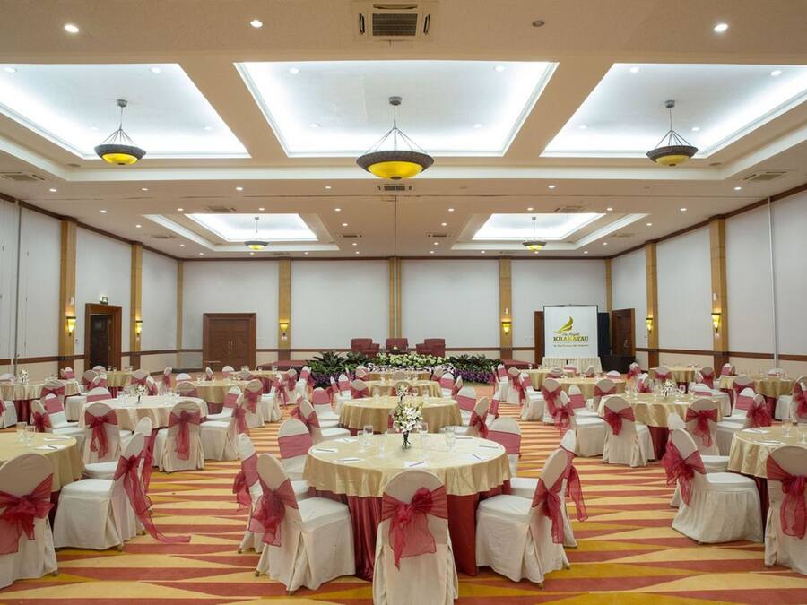 A large banquet hall with round tables, chairs, and a stage with floral arrangements.