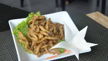 Un piatto quadrato bianco con cibo fritto, guarnito con lattuga e un fiore bianco, su un tavolo intrecciato.