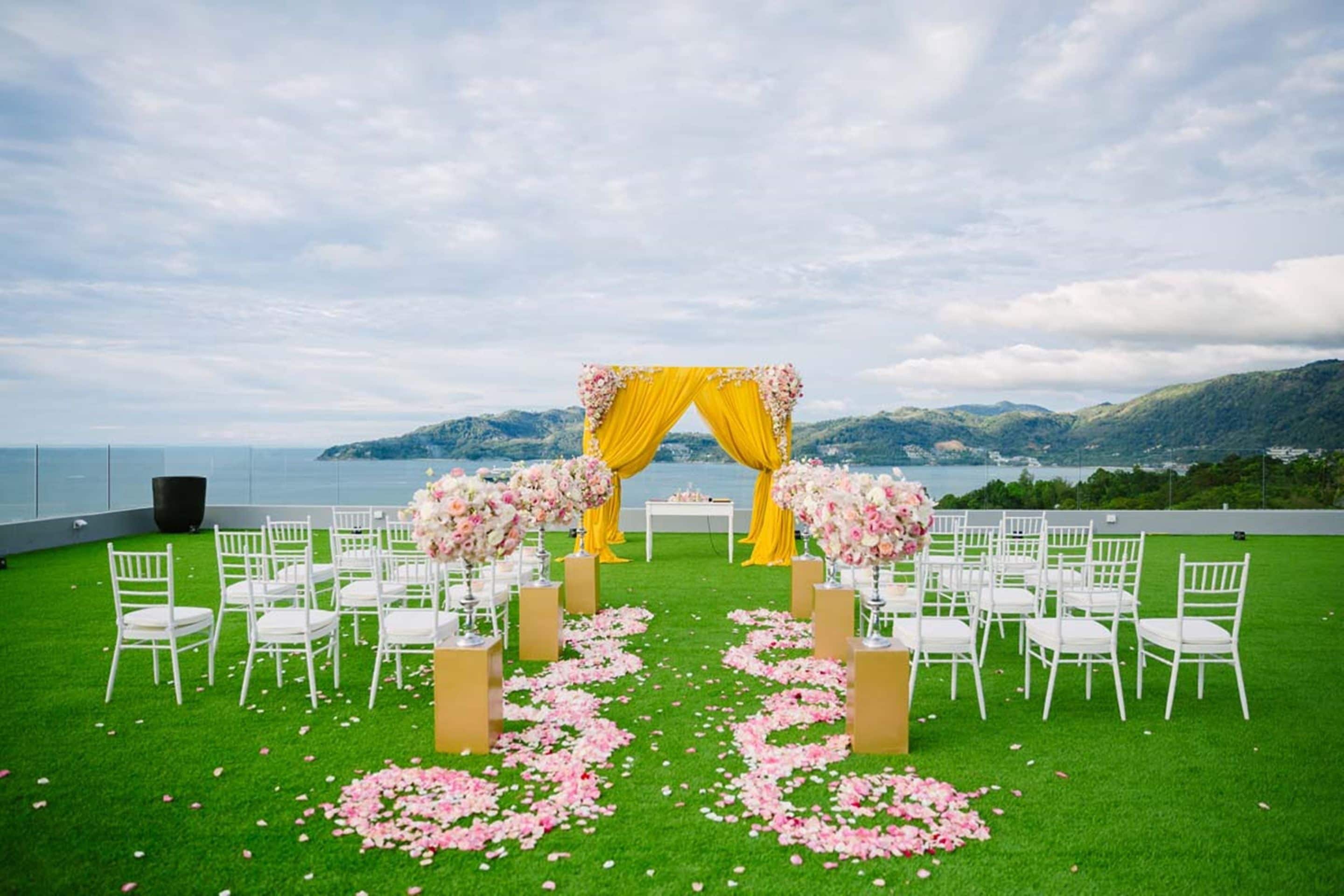 Une cérémonie de mariage en plein air, avec des chaises blanches disposées en rangées, une arche florale jaune et rose, et en arrière-plan, une vue sur un plan d’eau et des collines.