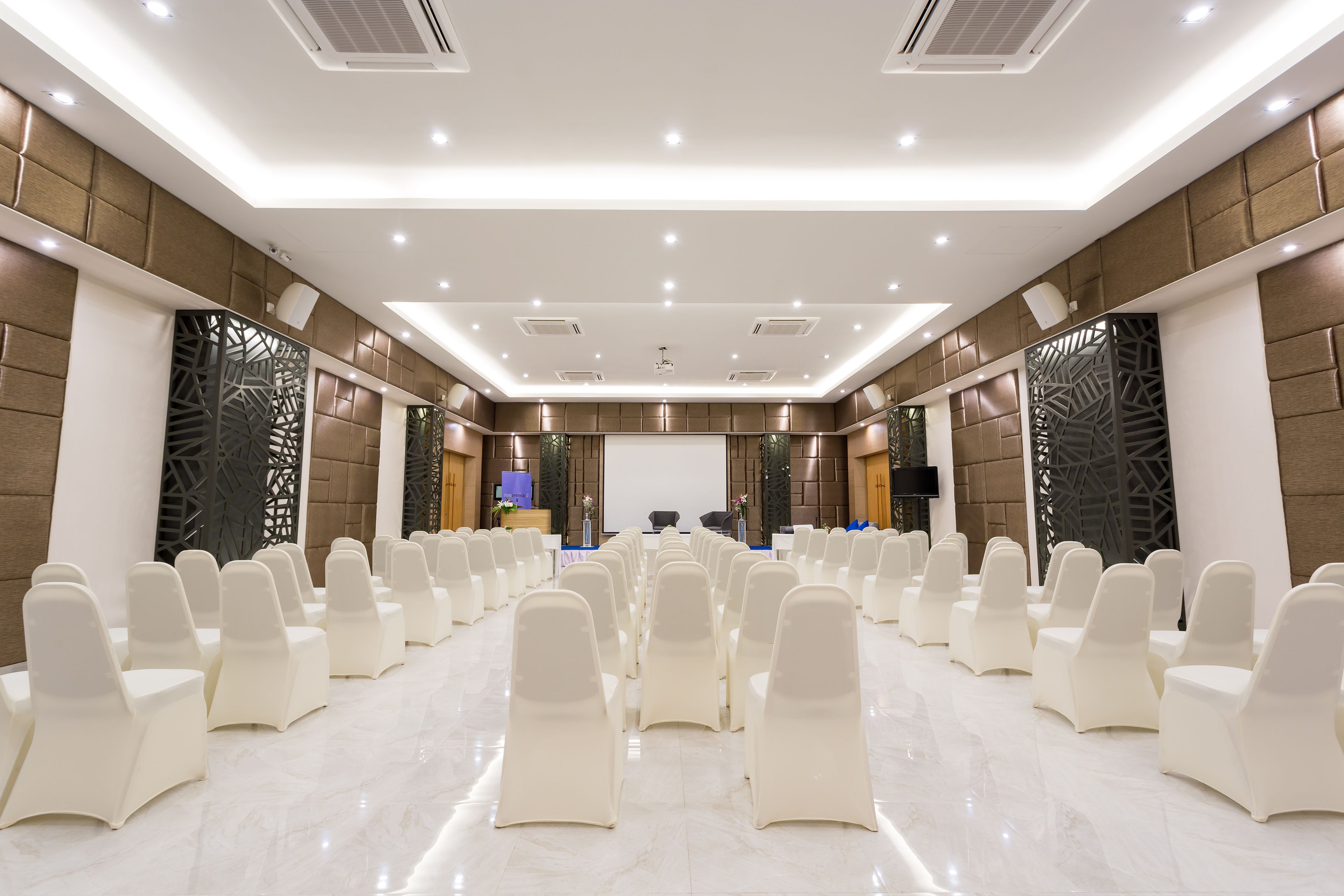 A conference room with rows of white chairs, a podium, and a large screen at the front.