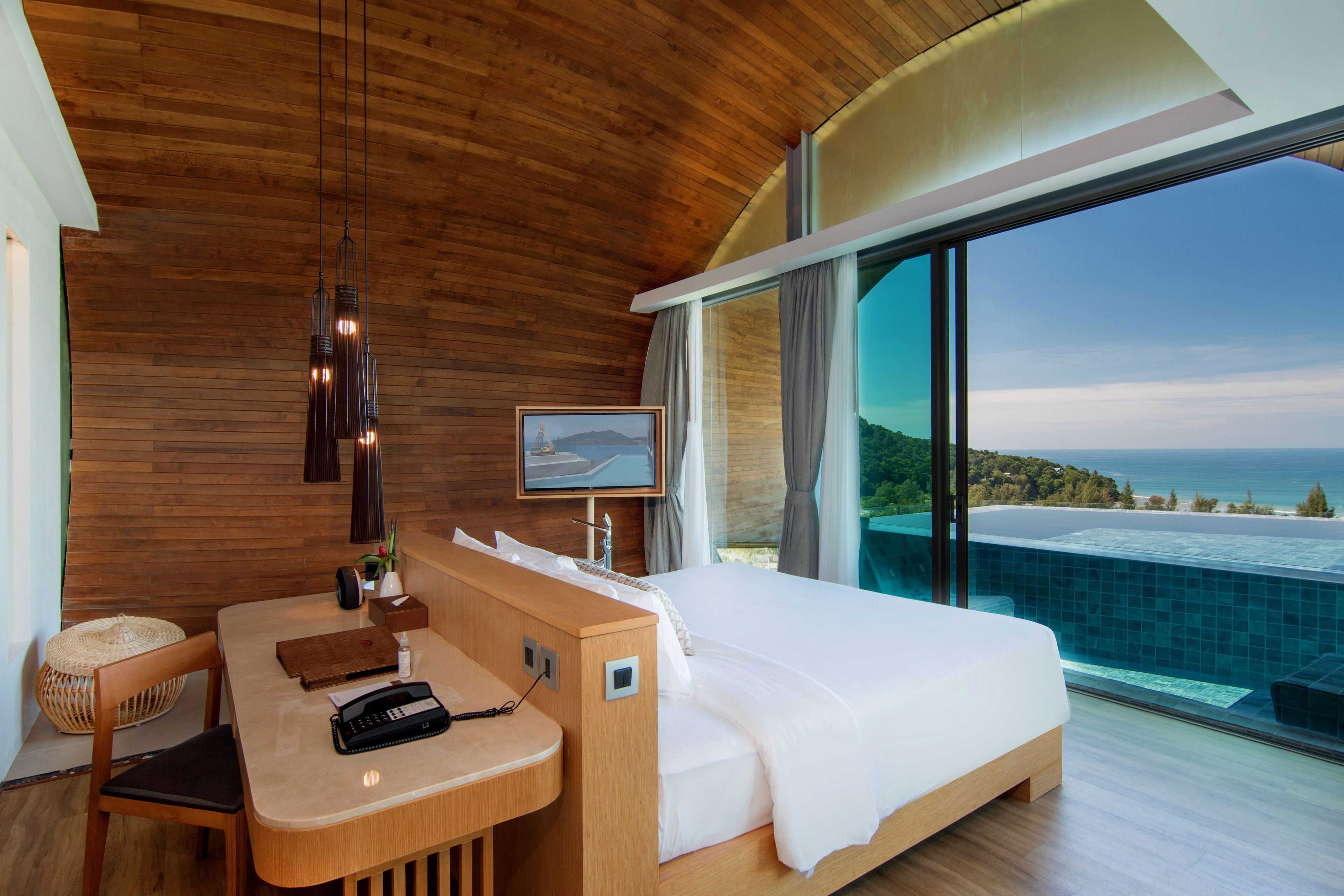 A modern hotel room with a wooden ceiling, a large bed, a desk with a telephone, and a view of the ocean.
