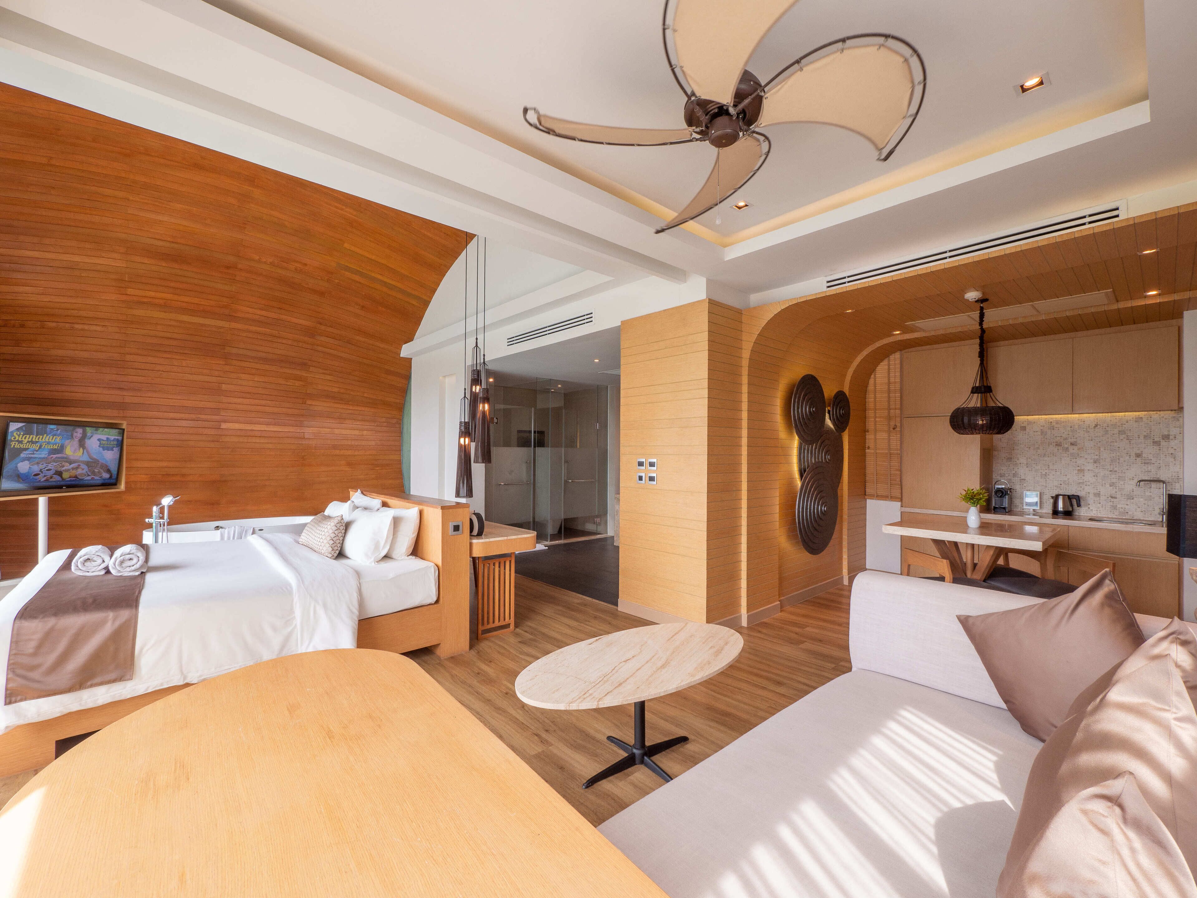 Ein modernes Hotelzimmer mit einer Wand in Holzoptik, einem Deckenventilator, einem Fernseher, einem Bett mit ordentlich gefalteten Handtüchern, einem runden Tisch und einer Küchenzeile.