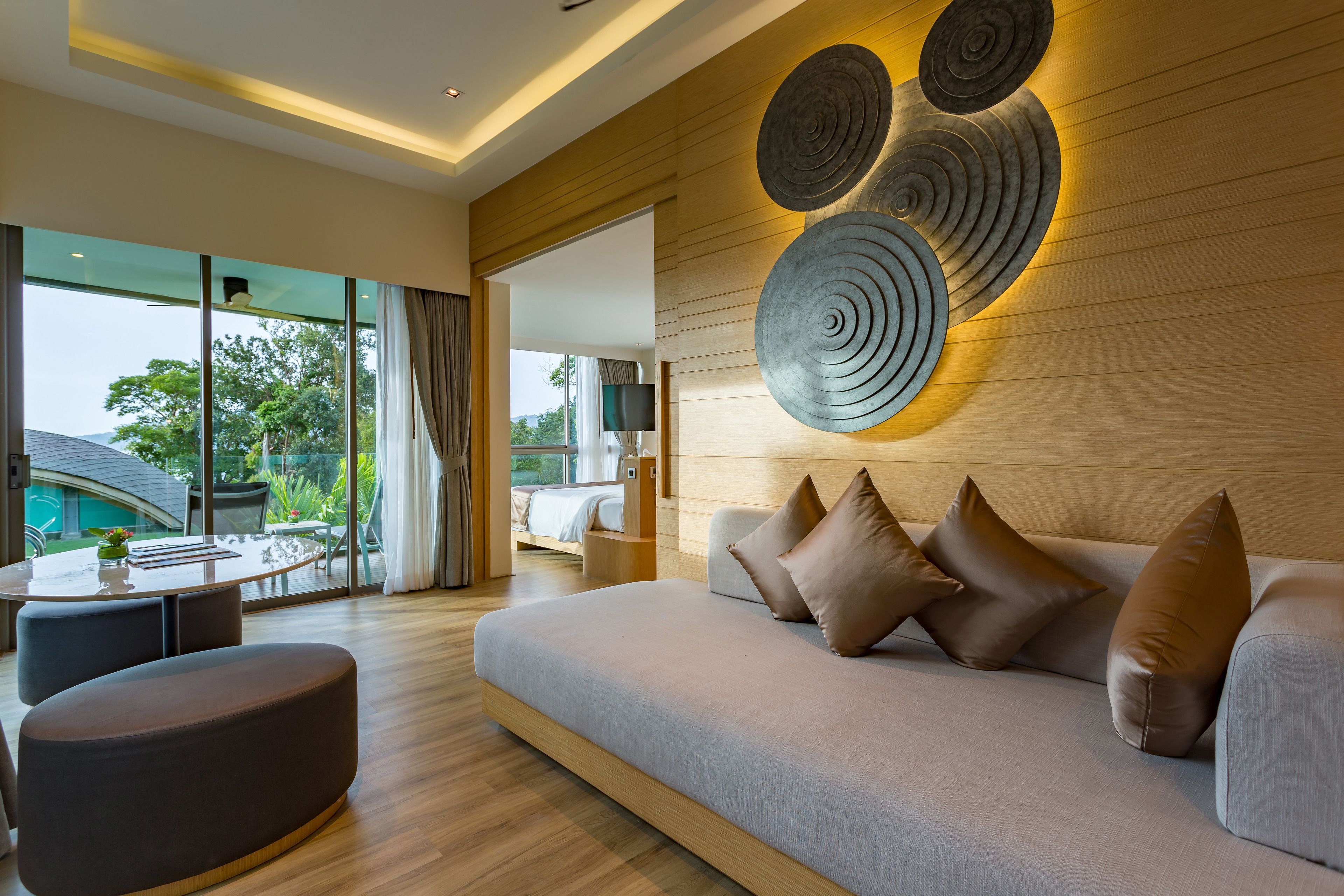A modern living room with a large sofa, a coffee table, and a TV. There is a wooden wall with circular decorations and a view of the outdoors.