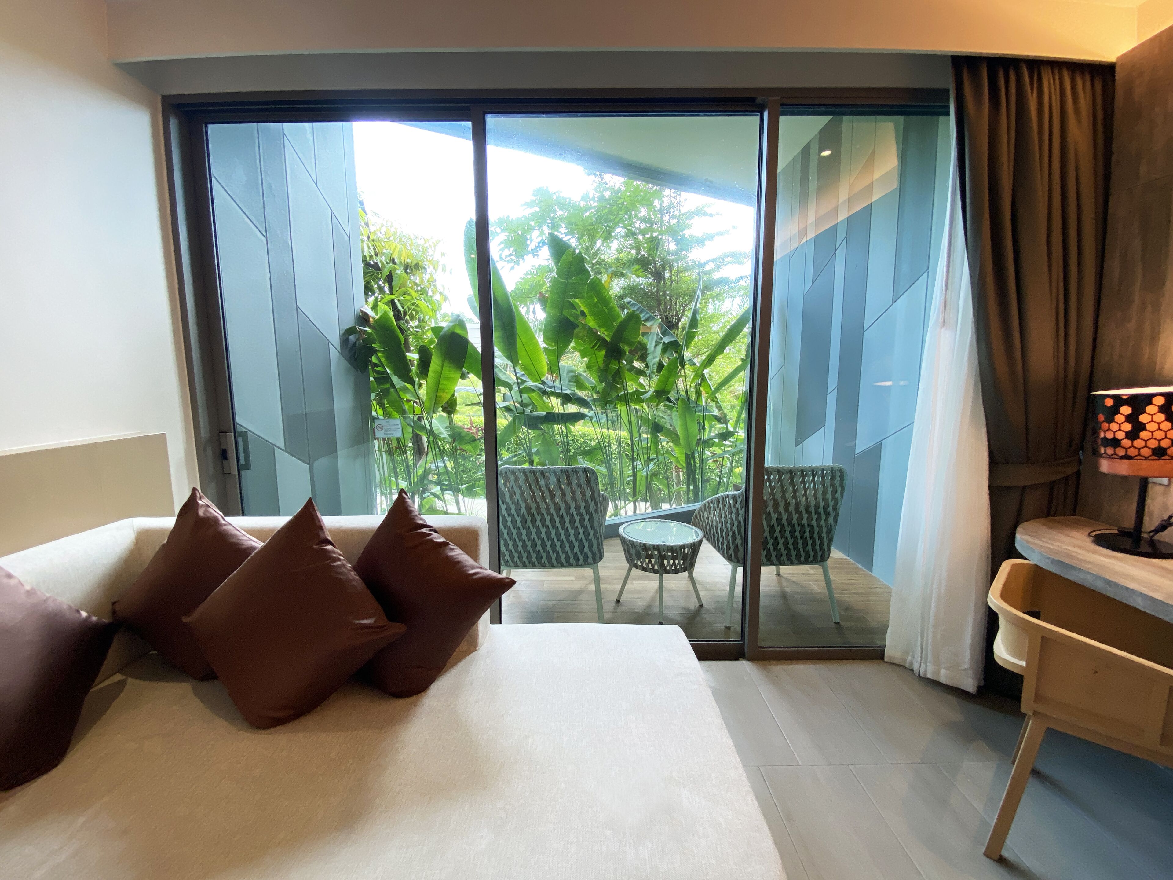 Habitación de hotel con cama, escritorio, silla y vista a un balcón con plantas.