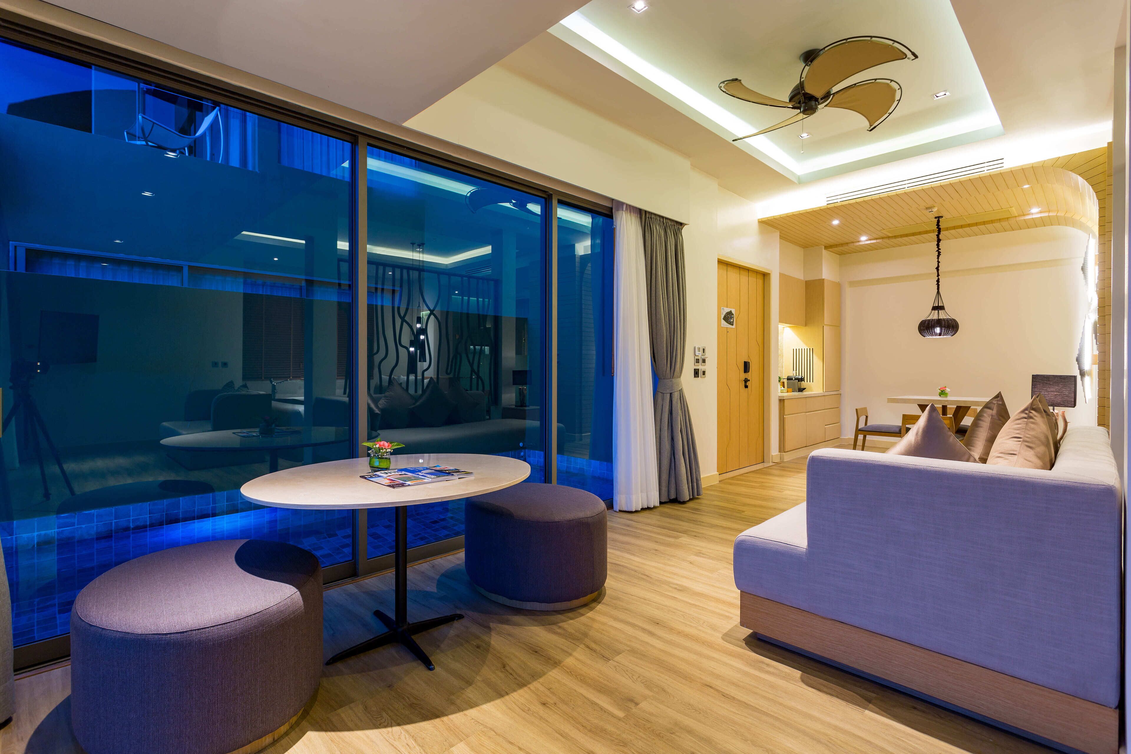 A modern hotel room with a large sliding glass door leading to a balcony, a round table with a chair, a sofa, and a ceiling fan.