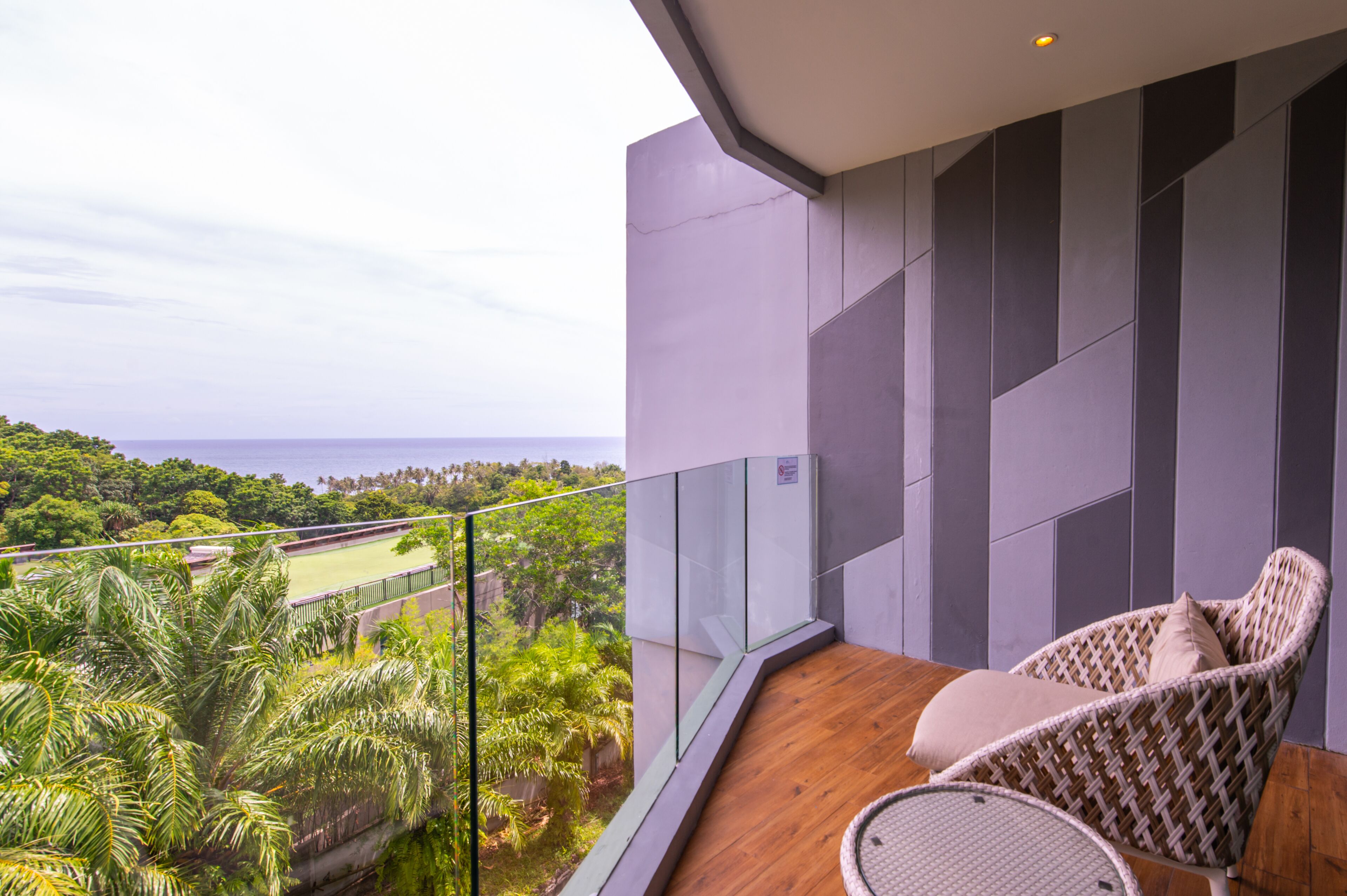 A balcony with a wicker chair and a small table, overlooking a lush green landscape and the sea.