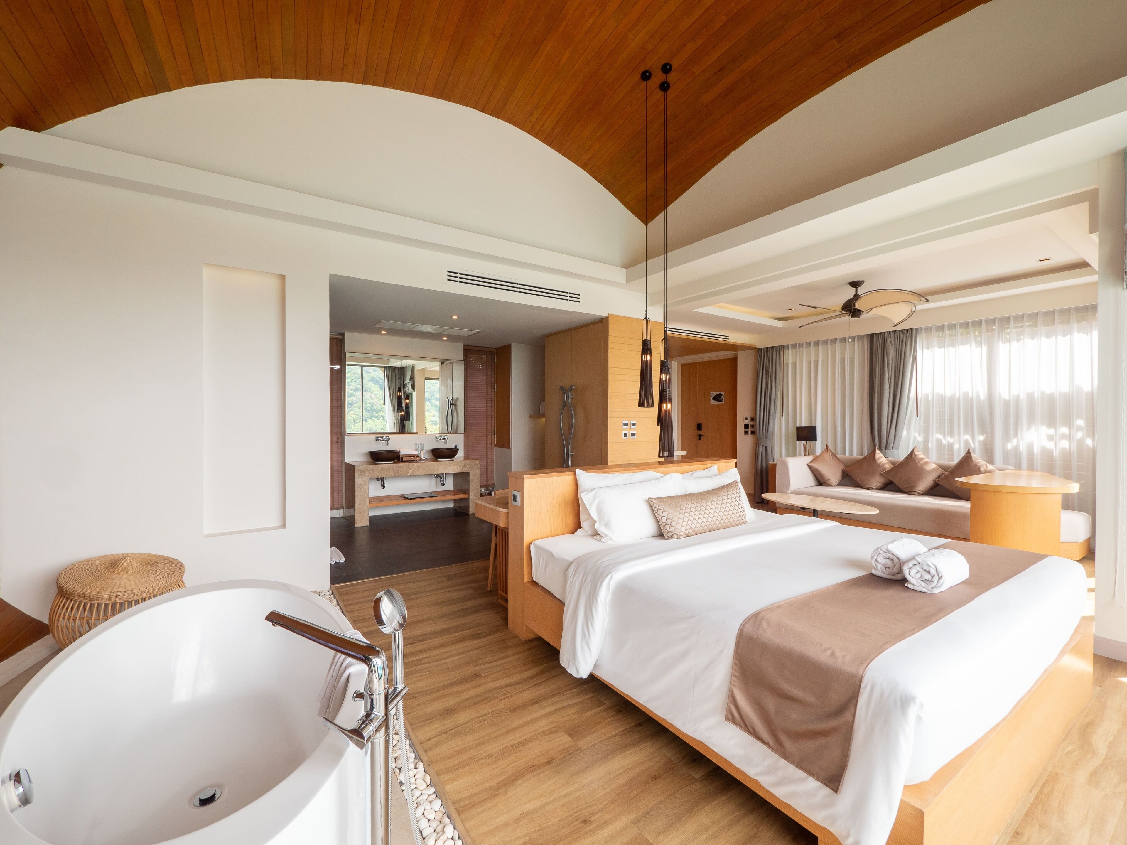 A modern hotel room with a large bed, a separate sitting area, a bathtub, and a large window with curtains.