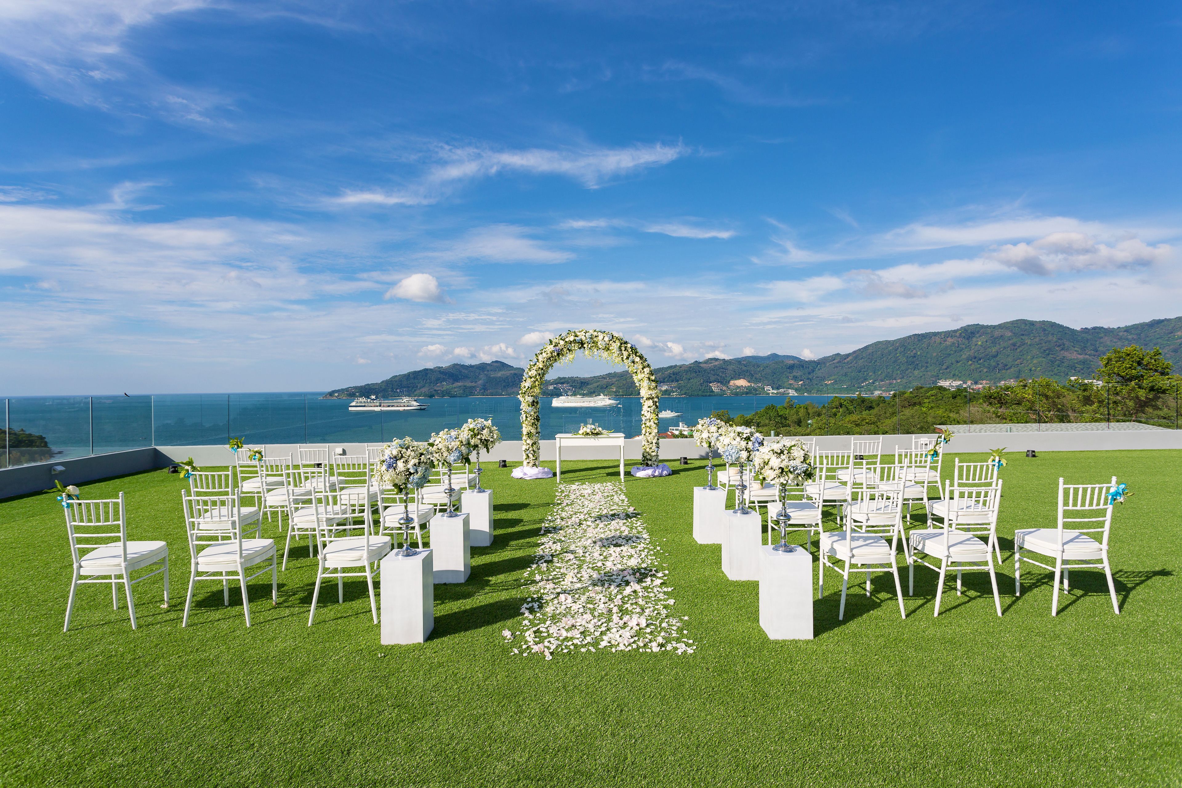 An outdoor wedding ceremony setup with white chairs and a floral arch, overlooking a coastal area.