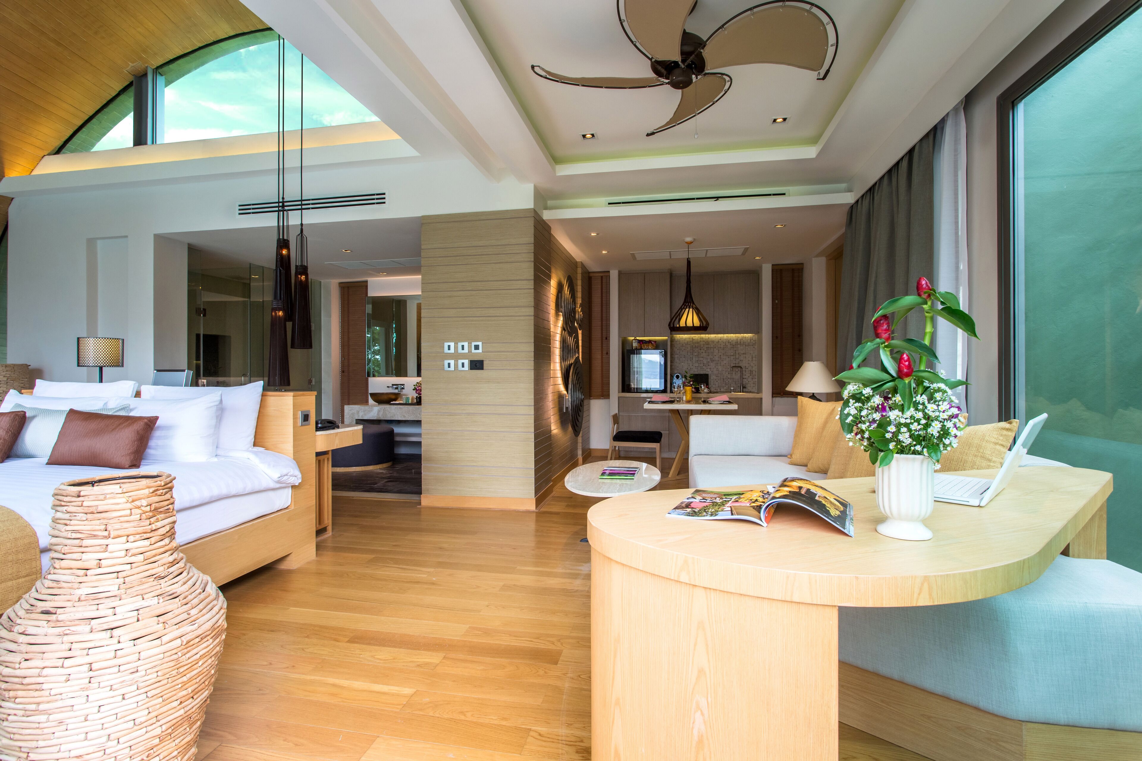 A modern hotel room with a large bed, a curved wooden desk, a laptop, and a dining area with a view.