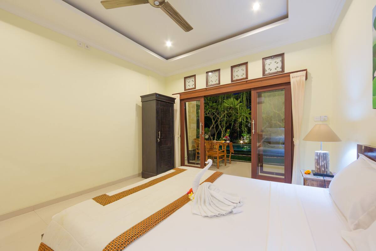 A hotel room with a bed, a ceiling fan, a desk with a lamp, and a view of a pool through a glass door.