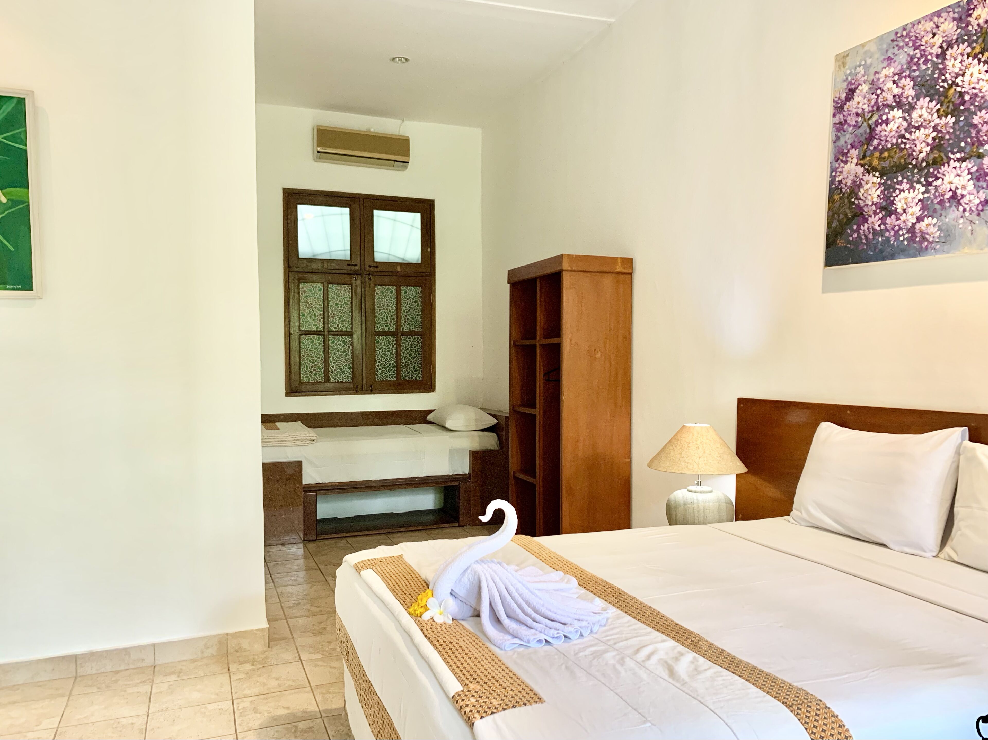 A hotel room with a bed, a wooden wardrobe, a window with a decorative grille, a wall-mounted air conditioner, and a painting of flowers.