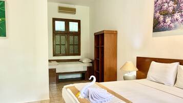 A hotel room with a bed, a wooden wardrobe, a window with a decorative grille, a wall-mounted air conditioner, and a painting of flowers.