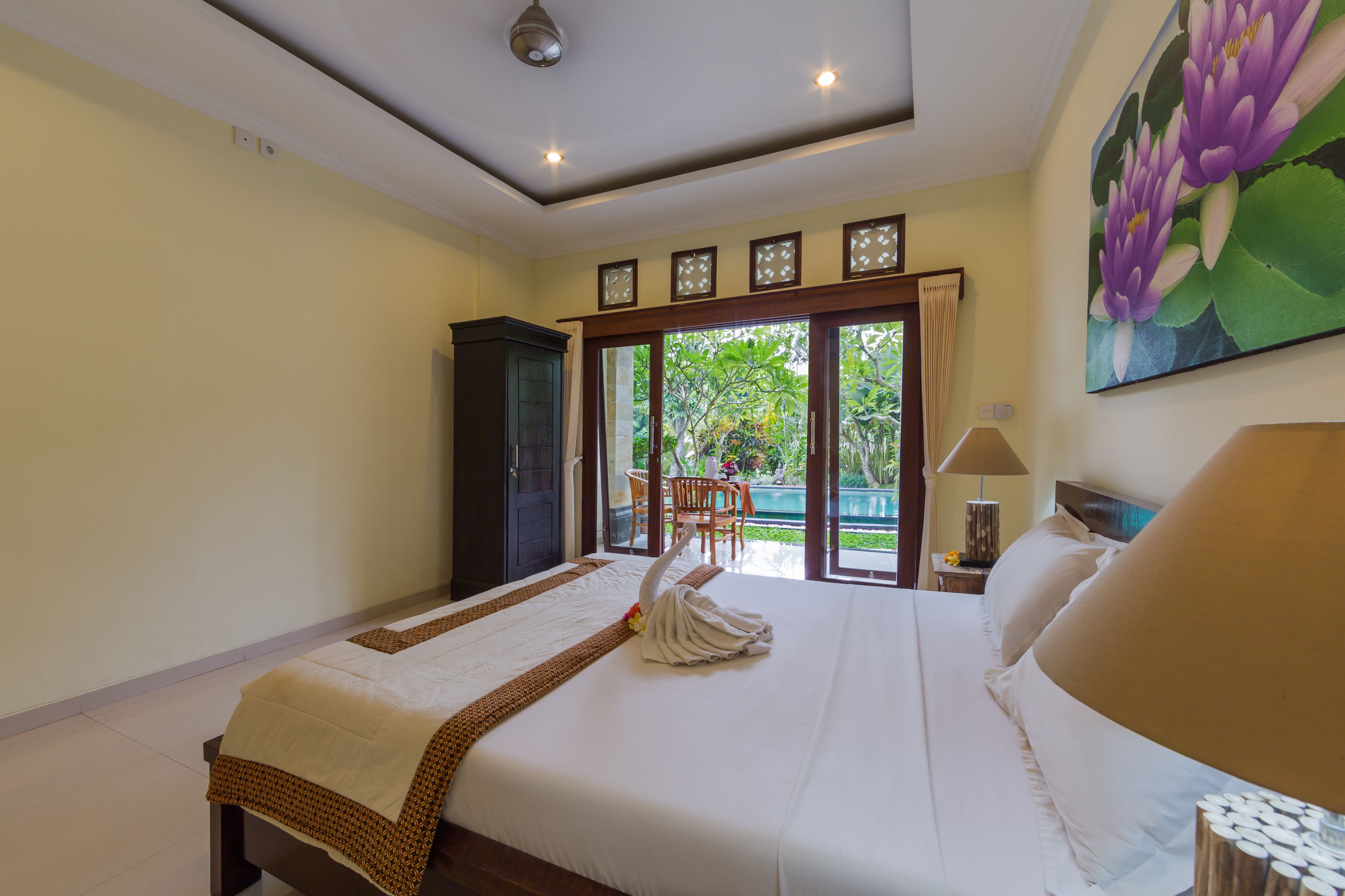 A hotel room with a bed, a wooden headboard, a painting of flowers, a window with a view of greenery, and a door leading to a balcony.