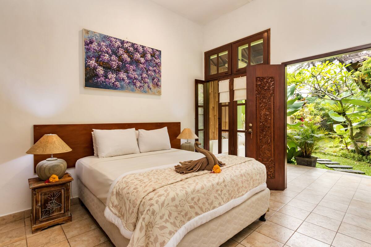 A bedroom with a large bed, a nightstand with a lamp, a painting on the wall, and a view of a garden through a glass door.
