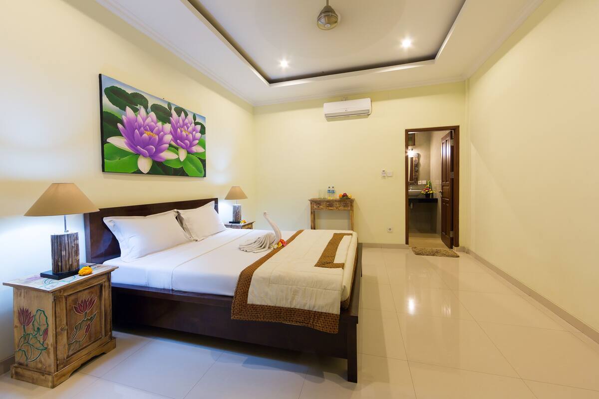 A neatly arranged bedroom with a bed, bedside lamps, a decorative cabinet, a painting, and a hallway leading to a bathroom.