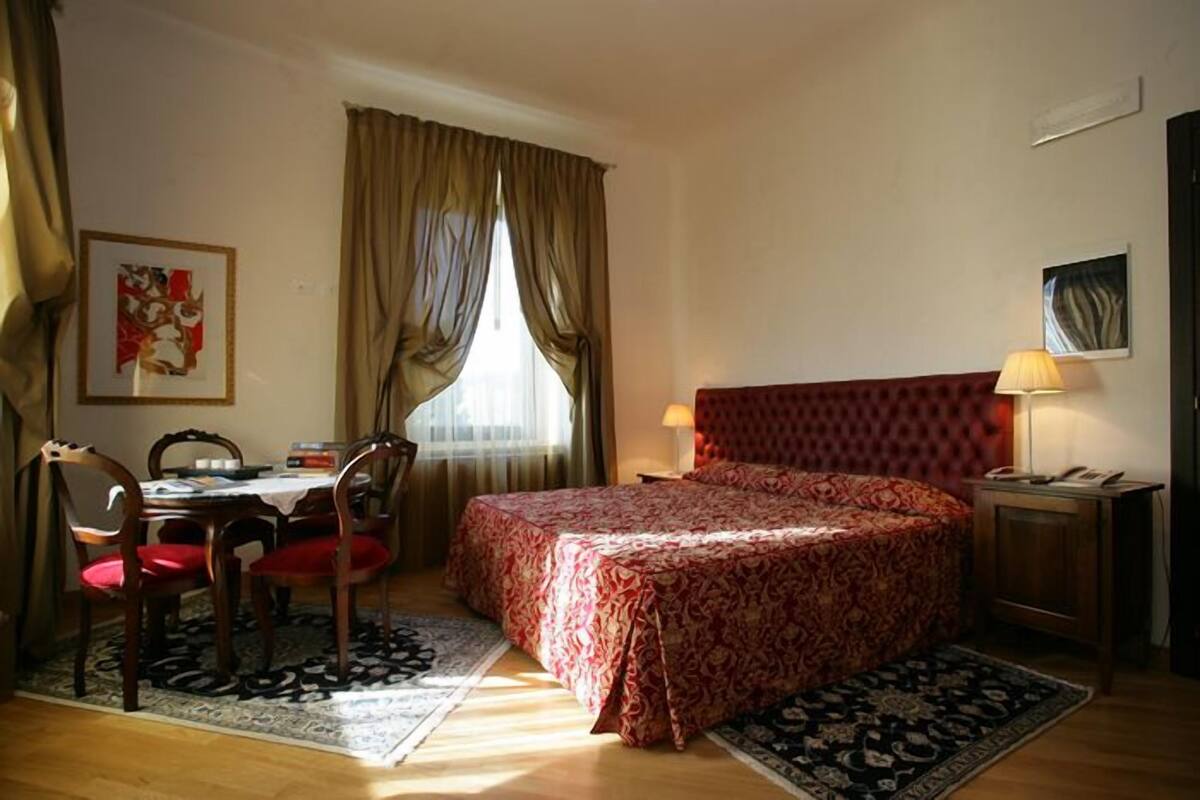 A hotel room with a bed, a desk, chairs, a lamp, and artwork on the walls.