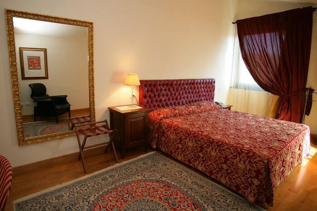 A bedroom with a large mirror, a bed with a red patterned bedspread, a small bedside table with a lamp, a chair, and a rug.