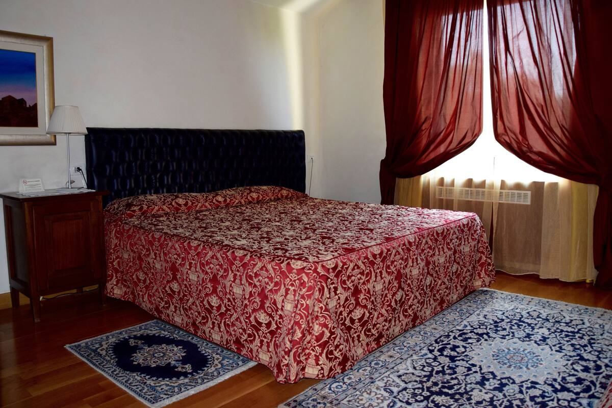 A bedroom with a bed, a nightstand, a lamp, a window with curtains, and a patterned rug.