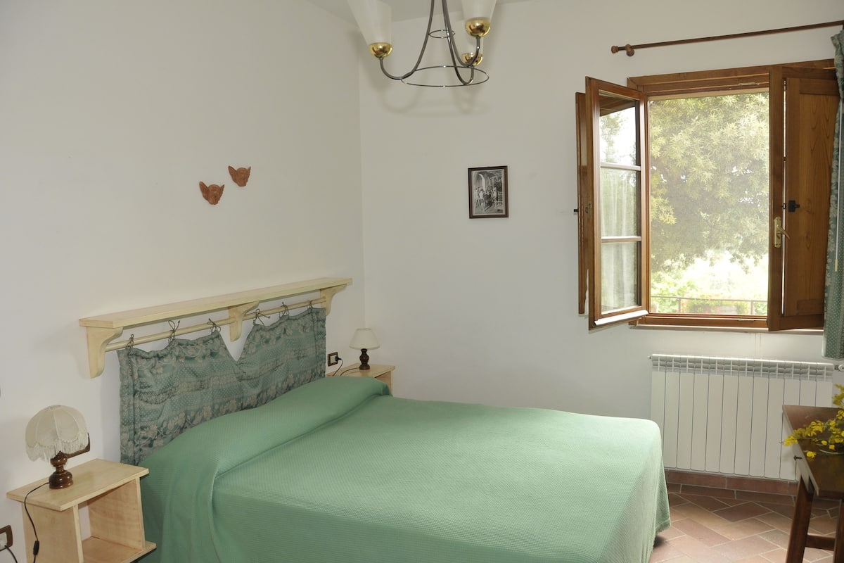 Un dormitorio con una cama, una mesita de noche, una lámpara, un radiador y una ventana con una persiana abierta.