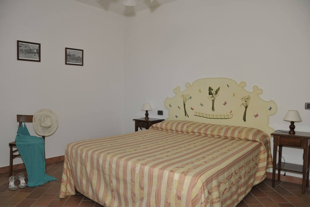 Un dormitorio con una cama, dos mesitas de noche, una silla con un ventilador y dos cuadros en la pared.