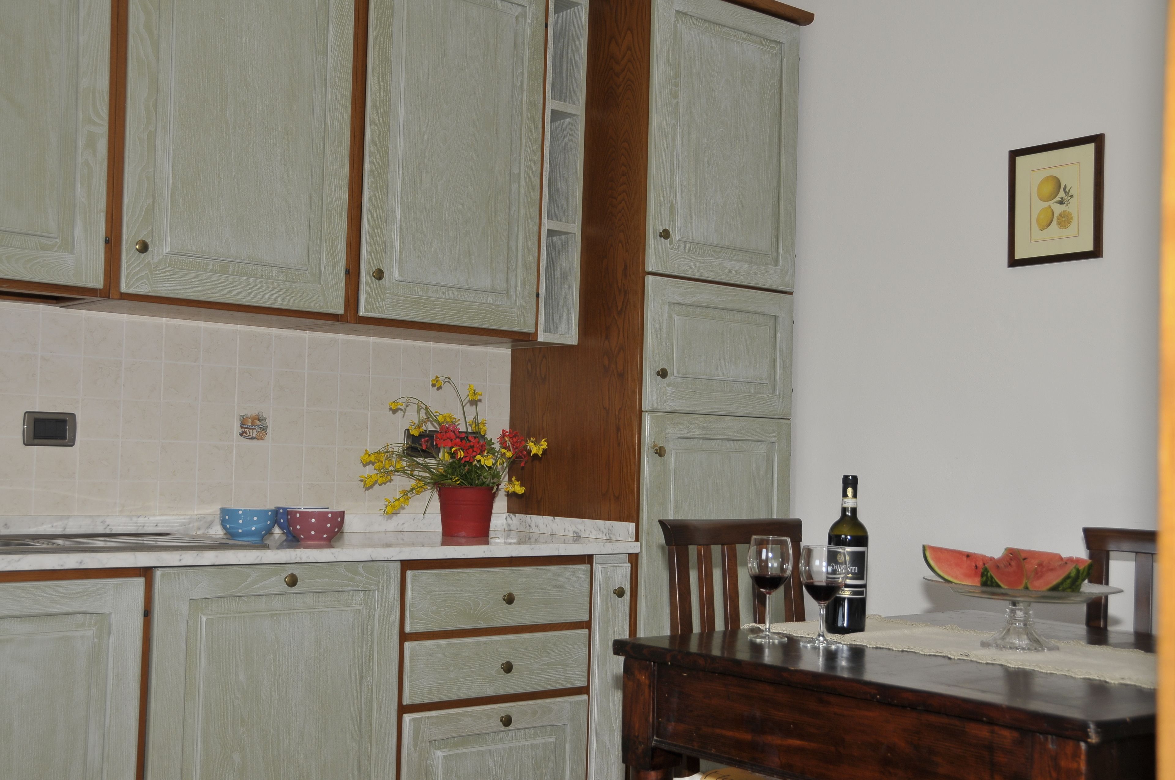 A kitchen with light green cabinets, a wooden chair, a dining table, a potted plant, a wine bottle, and a watermelon on a plate.