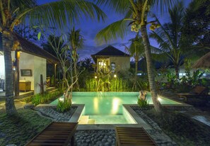 Ein Resort mit Swimmingpool, umgeben von Palmen und tropischer Vegetation, einer strohgedeckten Struktur und einem gut beleuchteten Eingang.
