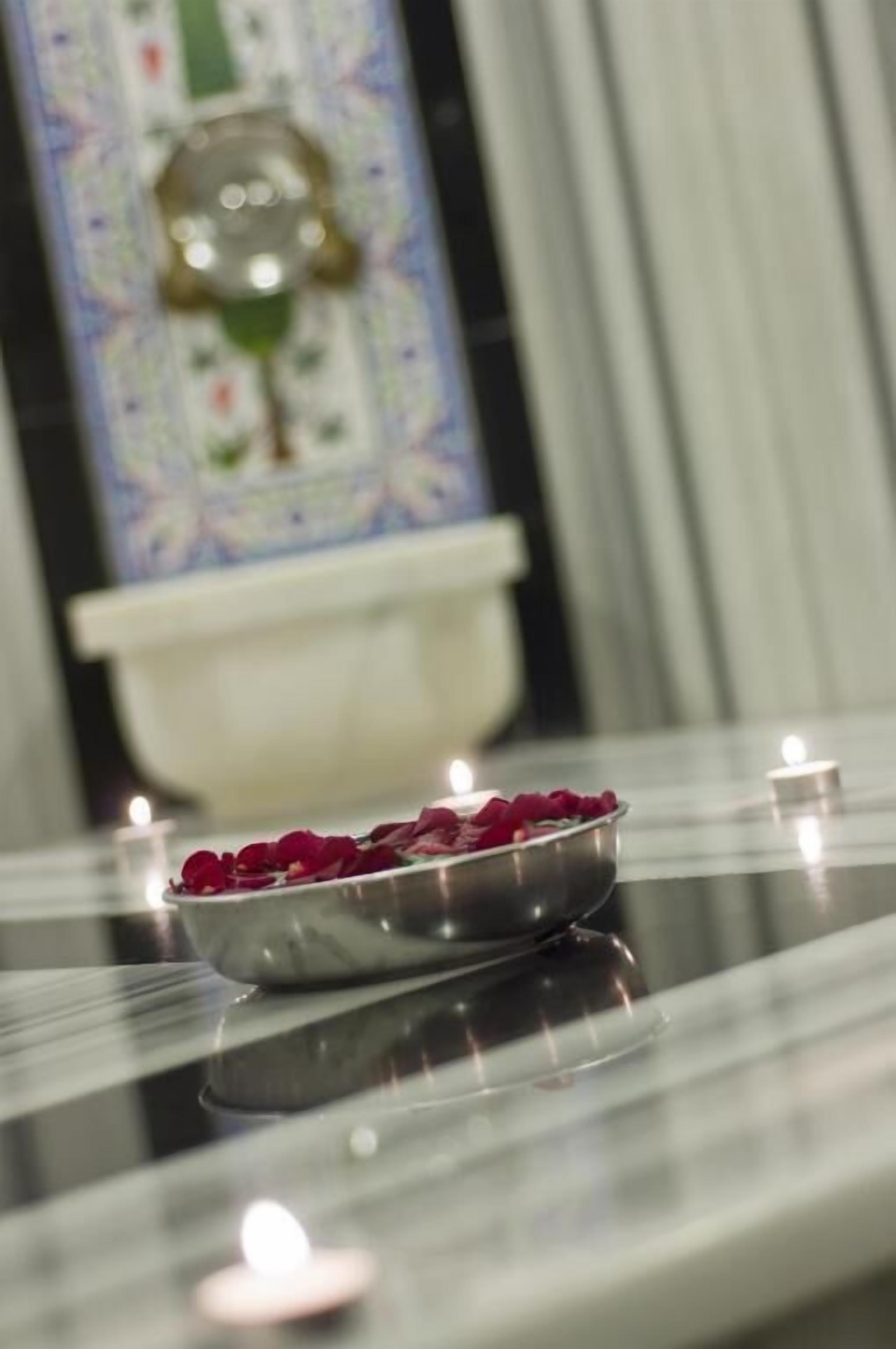 A polished table with a bowl of rose petals and lit candles, reflecting on the surface.