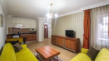 Un salon avec des canapés jaunes, une table basse en bois, un téléviseur posé sur un meuble en bois, et une cuisine à l’arrière-plan.