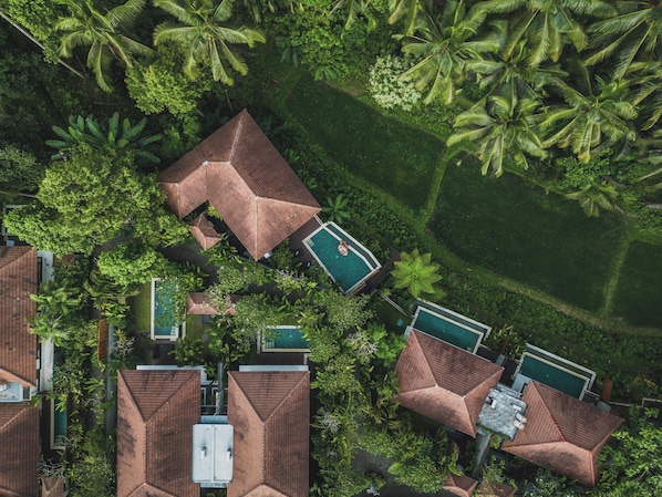 Vista aérea de un complejo turístico con varias construcciones, cada una con un diseño geométrico único en su techo, rodeado de exuberante vegetación y palmeras.