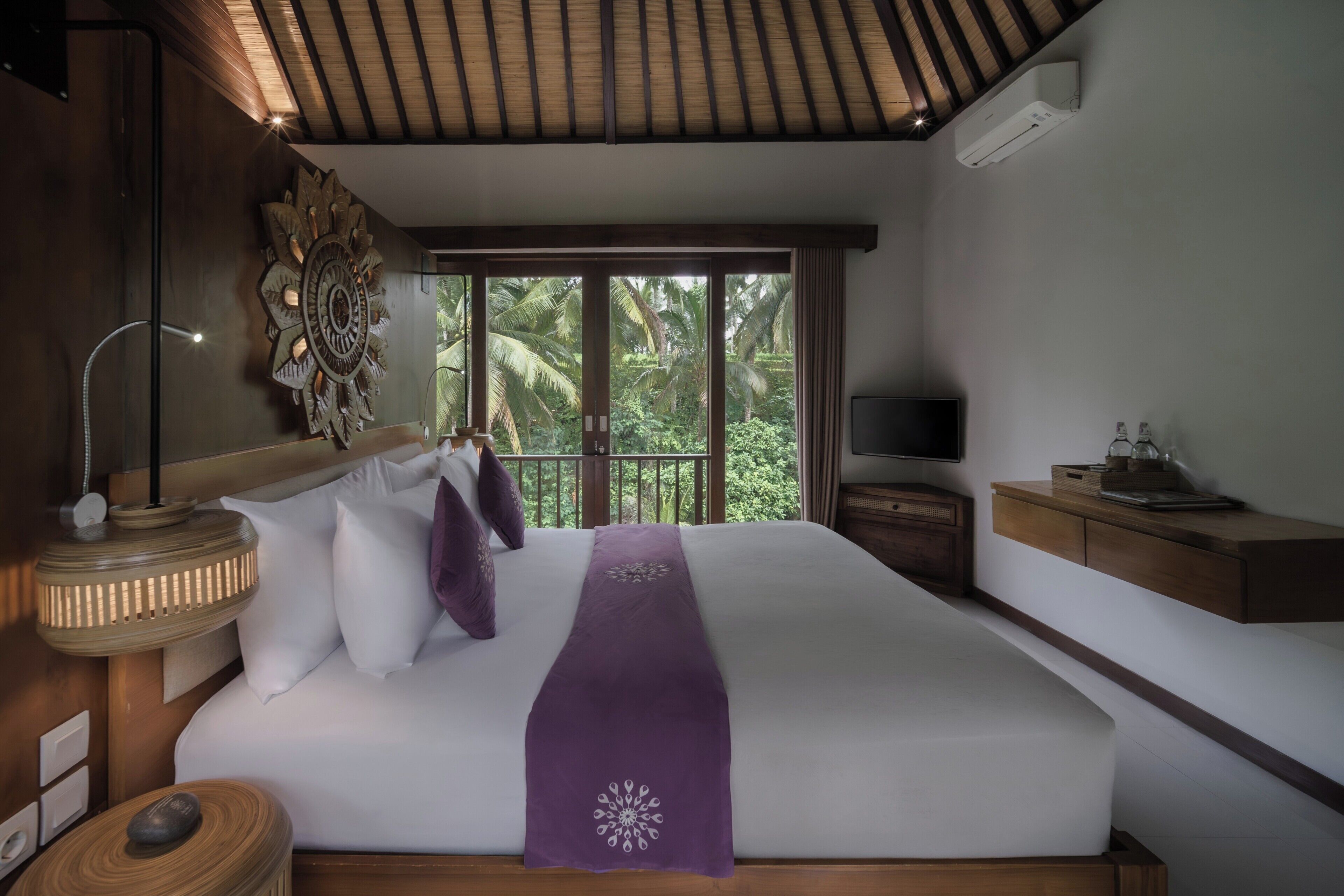 A bedroom with a bed, a TV, a nightstand, and a window with a view of greenery.
