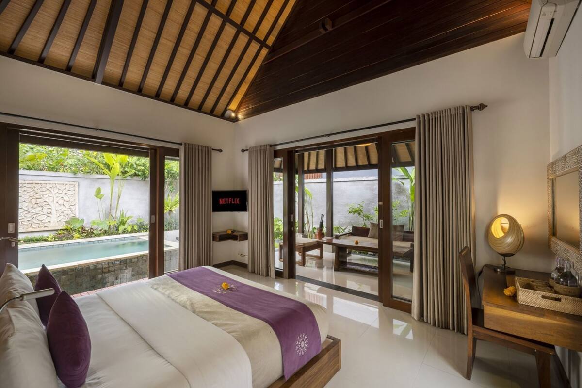 A bedroom with a bed, a TV, a desk with a lamp, and a view of a pool and greenery.