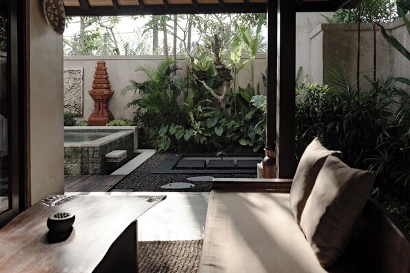A modern interior with a sofa, a small table, and a view of an outdoor garden area with plants and a decorative structure. - Dedary Resort & Spa Ubud by Ini Vie Hospitality (Tegallalang)