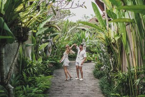 A pathway flanked by lush greenery and a statue, leading to a building with a tiled roof.
