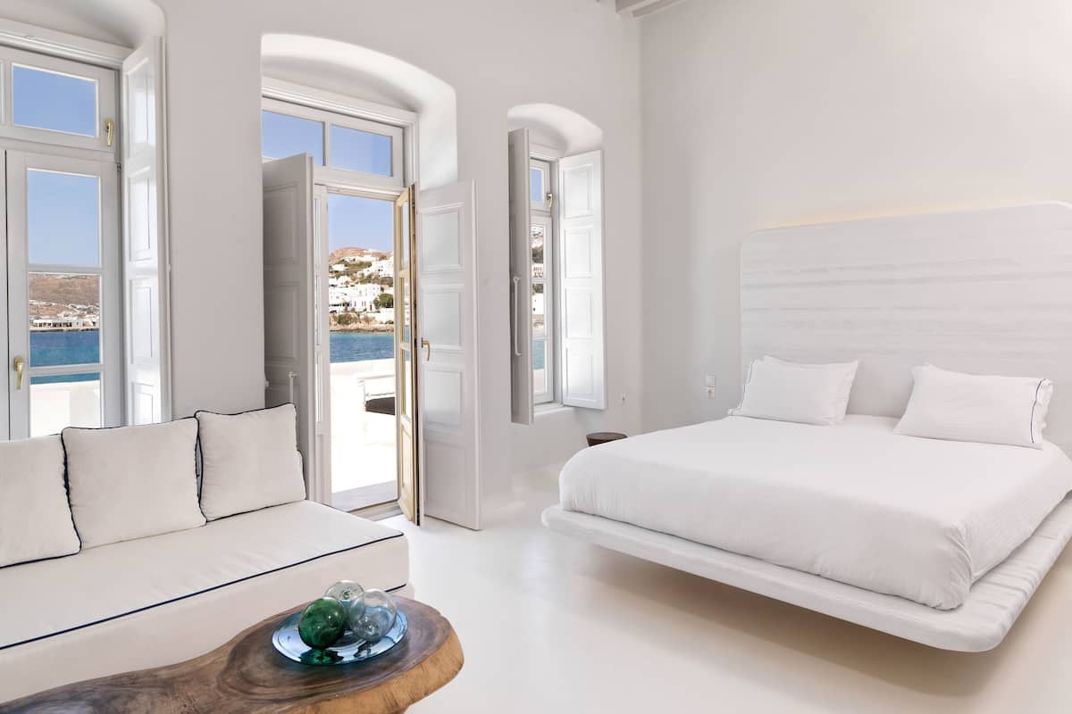 A modern bedroom with a large bed, a sofa, and a view of the sea through open doors.