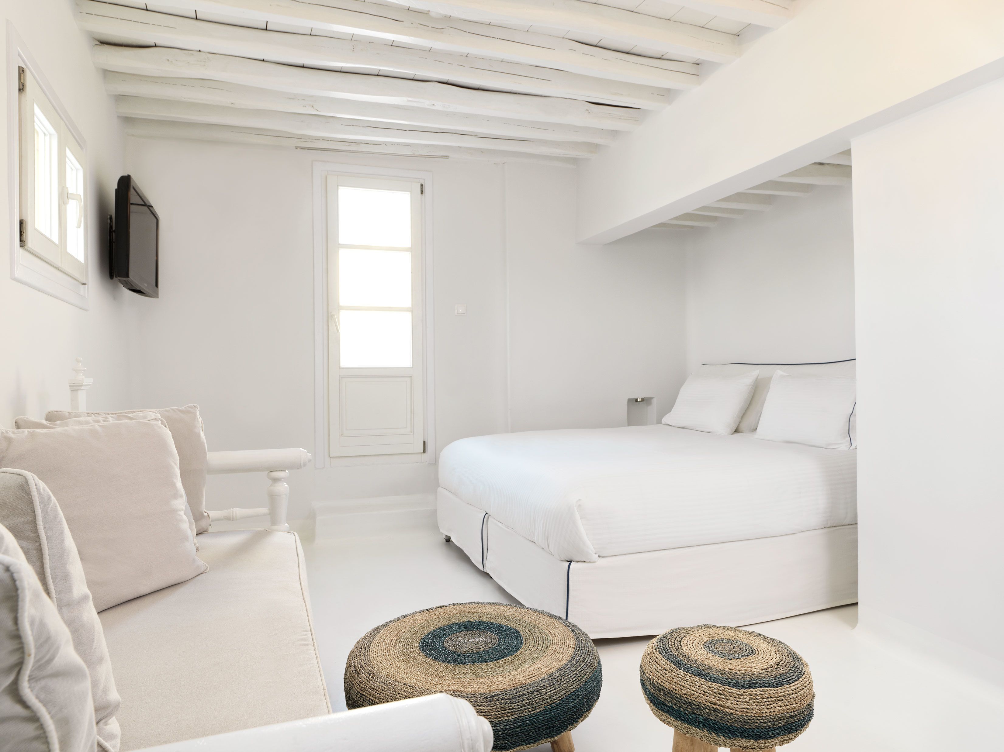 A minimalist white bedroom with a bed, a small white table, and two woven stools.