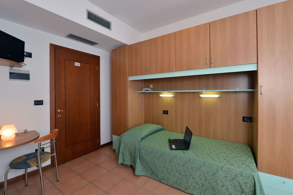 Ein kleiner, hell erleuchteter Raum mit einem Bett, einem Schreibtisch mit Stuhl und einem an der Wand befestigten Fernseher.